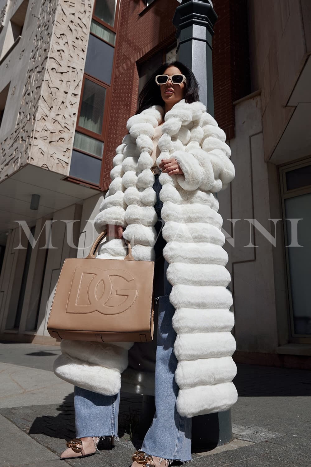 A woman wearing a chic long white faux fur coat, highlighting its high-quality, fluffy fabric and a coordinating belt that accentuates her figure. The coat’s luxurious design makes it a stunning choice for formal winter events.