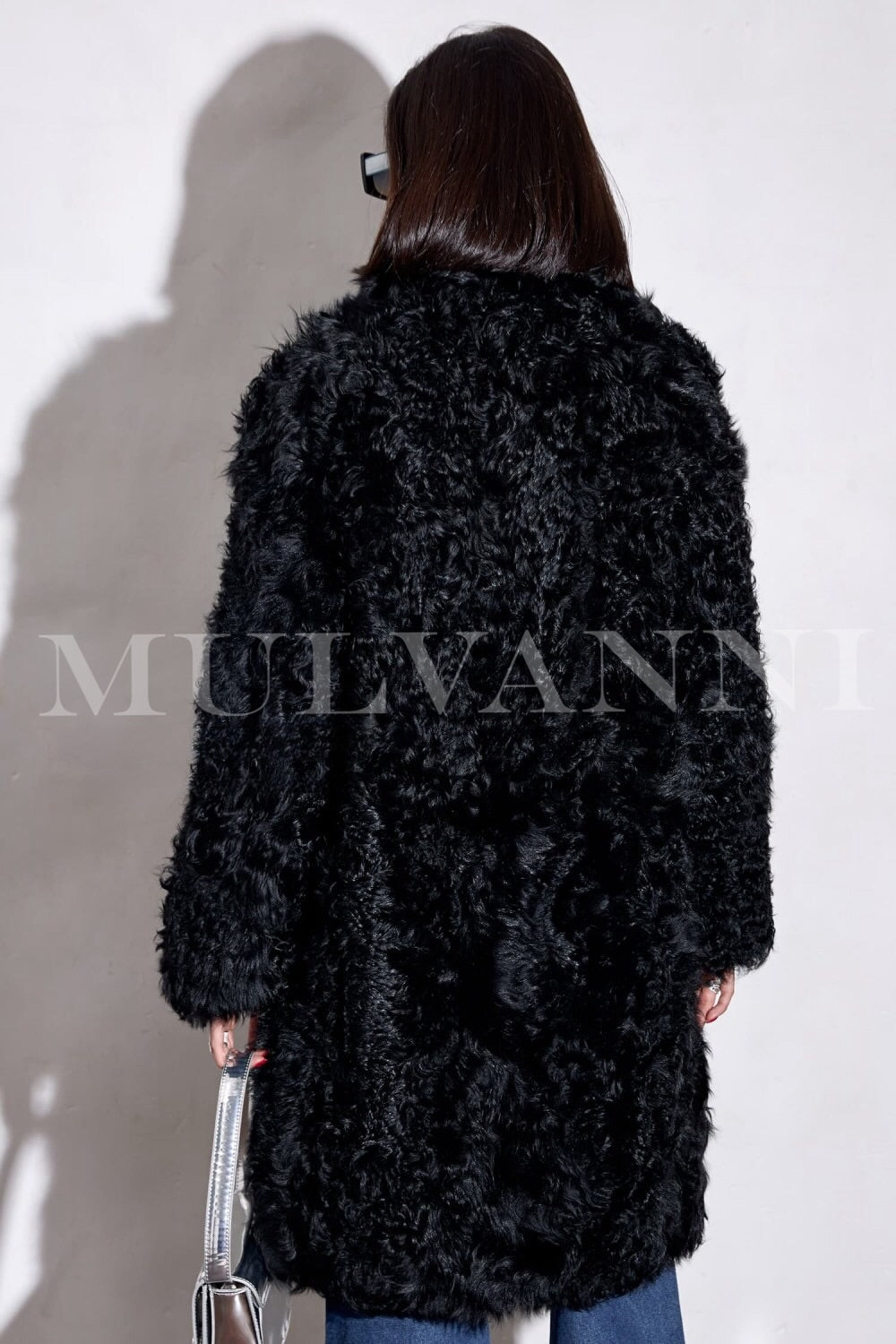 A woman wearing a striking long black shearling coat, featuring a plush texture and a flattering silhouette. Her stylish sunglasses complete the look, adding an air of sophistication to her winter outfit.