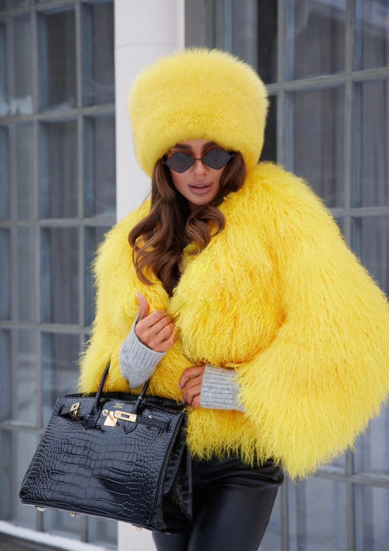 A woman wearing a bright yellow fox fur hat along with a coordinating Mongolian fur coat. The hat's plush texture contrasts beautifully with the coat's luxurious fur, creating a fashionable and eye-catching winter outfit.