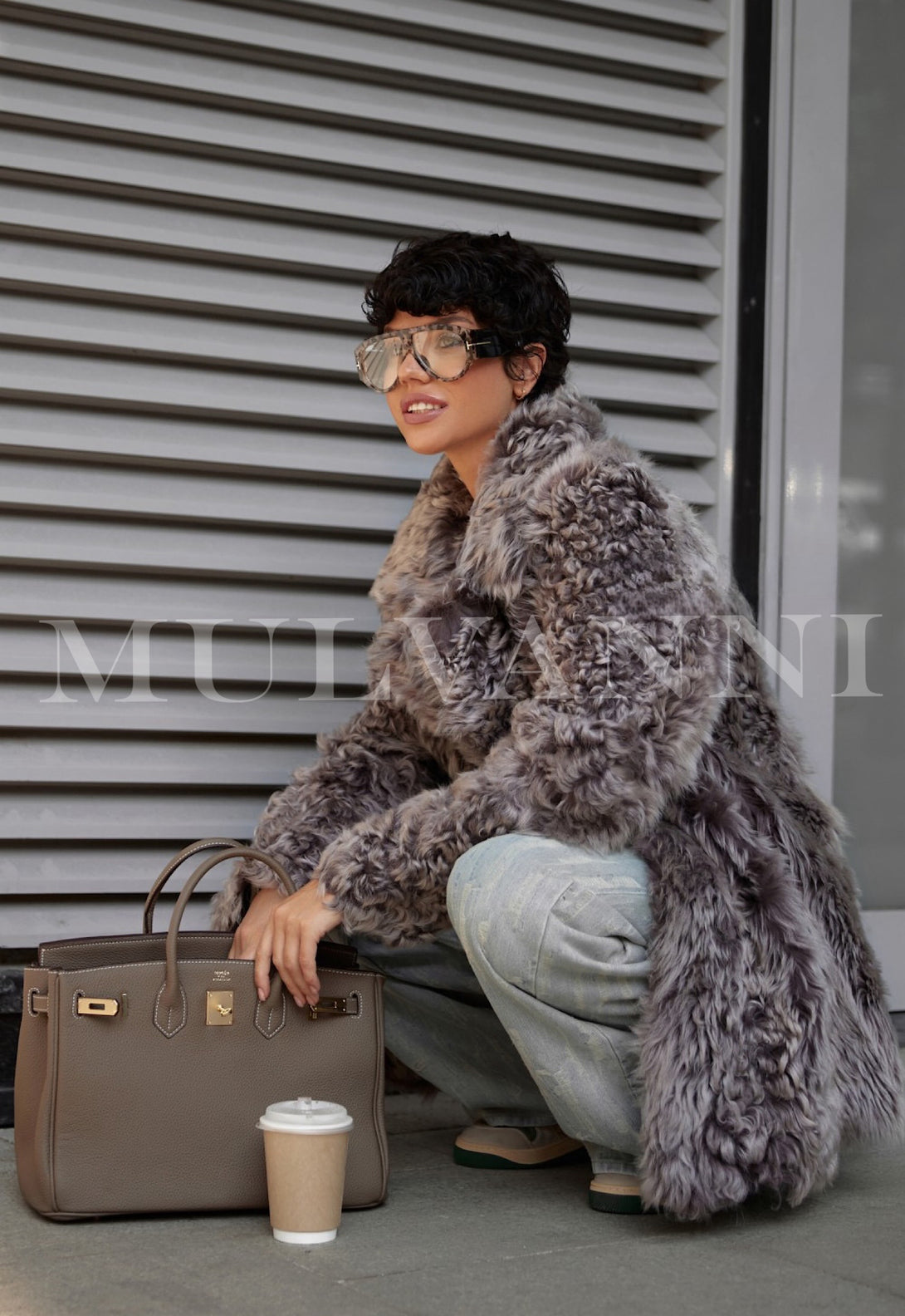 Woman wearing luxurious ADRIE mousse shearling coat, sitting outdoors with grey coat, handbag, and coffee cup, epitomizing elegant fashion.