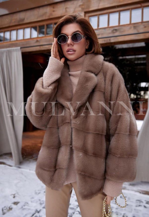 A woman wearing a short brown mink jacket paired with stylish sunglasses. The jacket features a luxurious, soft texture, adding a touch of elegance to her fashionable and chic look.