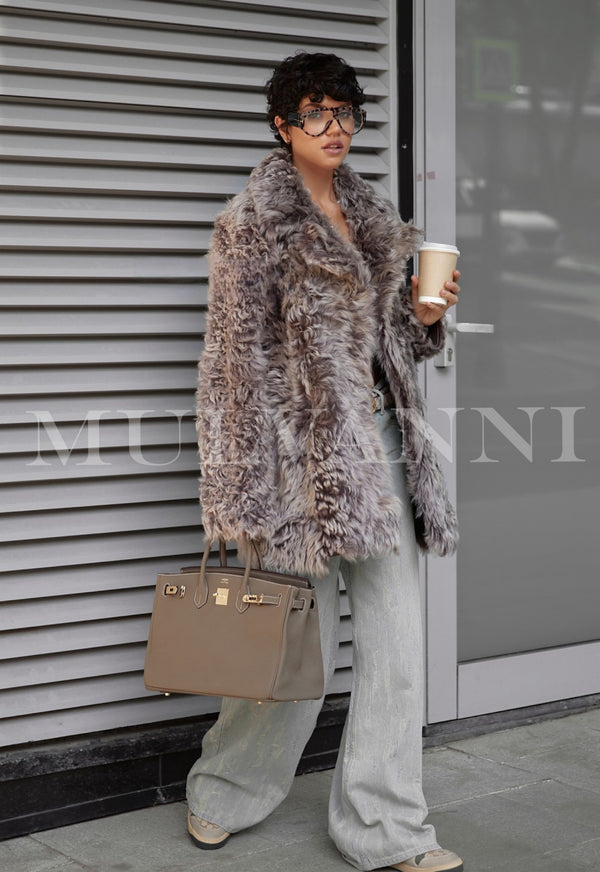 Woman wearing ADRIE Mousse Shearling Coat holding coffee and handbag standing in front of gray wall