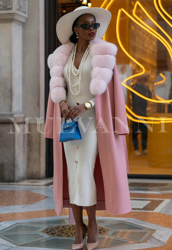 Woman wearing a luxurious cashmere coat with fox fur collar in blush pink, accessorized with a white dress, hat, pearls, and blue handbag.