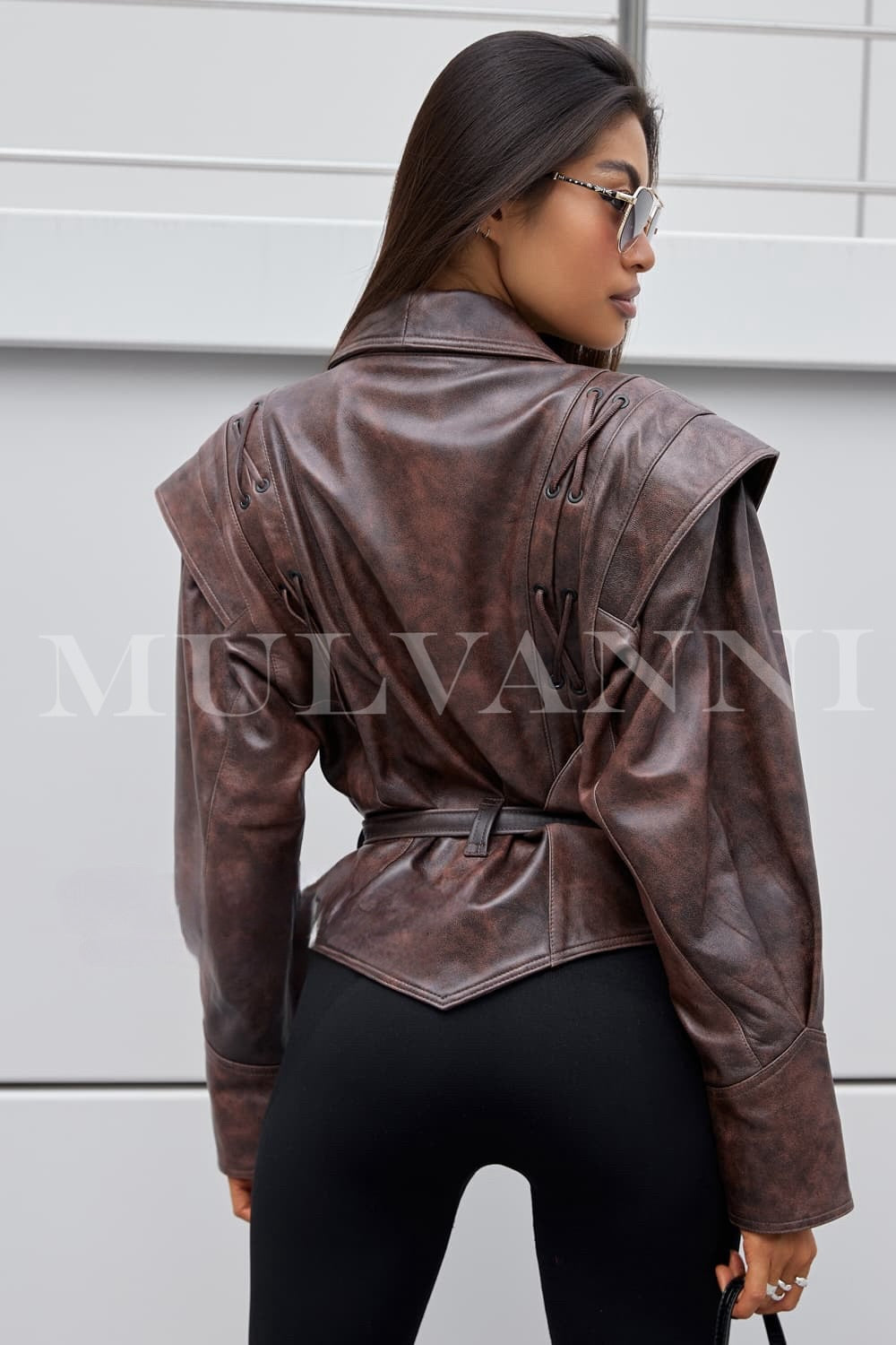 A woman dressed in a chic brown leather jacket, featuring a modern design and a matching belt that accentuates her waist. The jacket's sleek lines and rich texture create a fashionable statement piece.