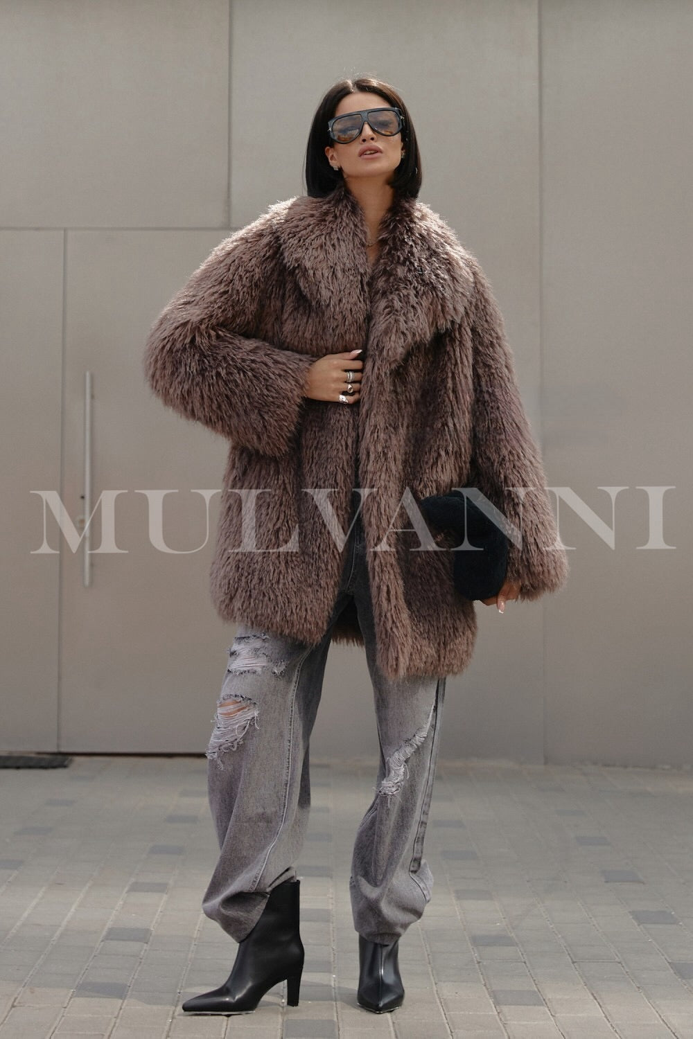 A woman dressed in a fashionable brown faux Mongolian coat, featuring a plush exterior and stylish sunglasses. The coat’s luxurious look and relaxed fit create an effortlessly chic winter outfit.