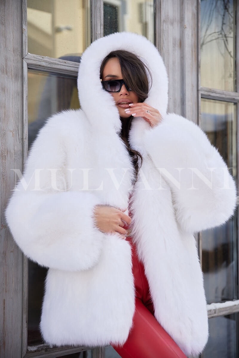 A woman wearing a luxurious white hooded fox fur coat, featuring a soft, fluffy texture and a stylish hood. The coat has a relaxed fit and falls to mid-thigh, exuding elegance and warmth for cold weather.