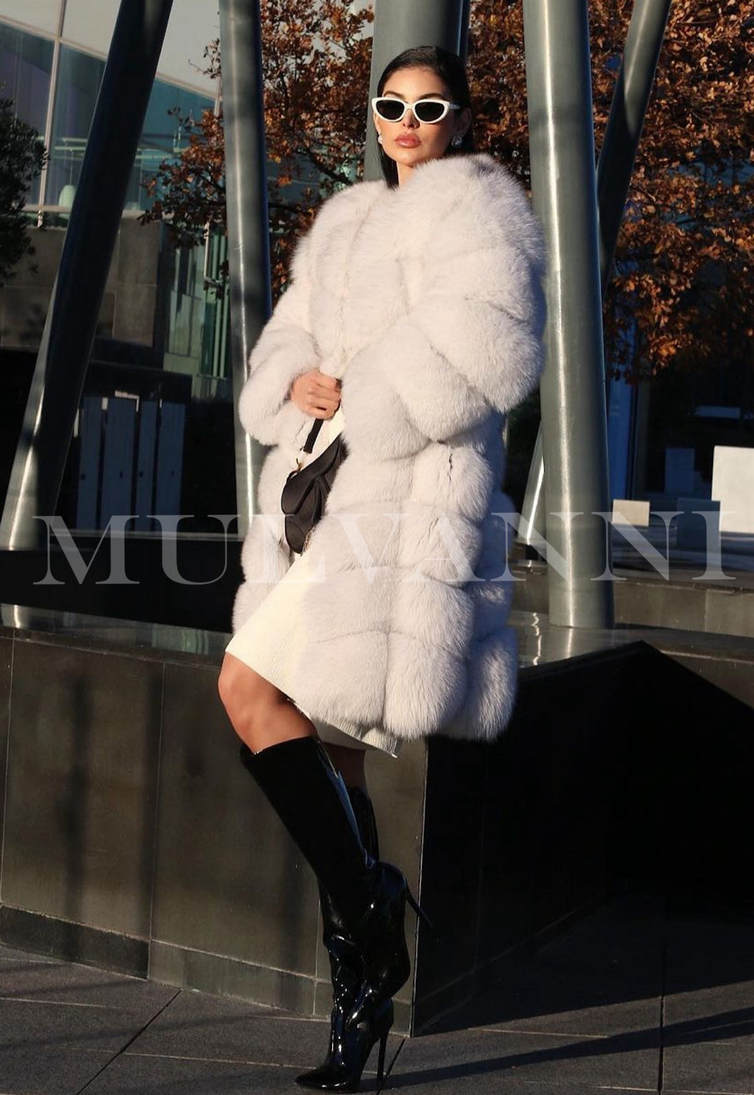 A woman dressed in a chic midi-length white fox fur coat, showcasing its soft and luxurious texture. The coat’s elegant design adds a touch of sophistication to her winter outfit paired with long boots. 