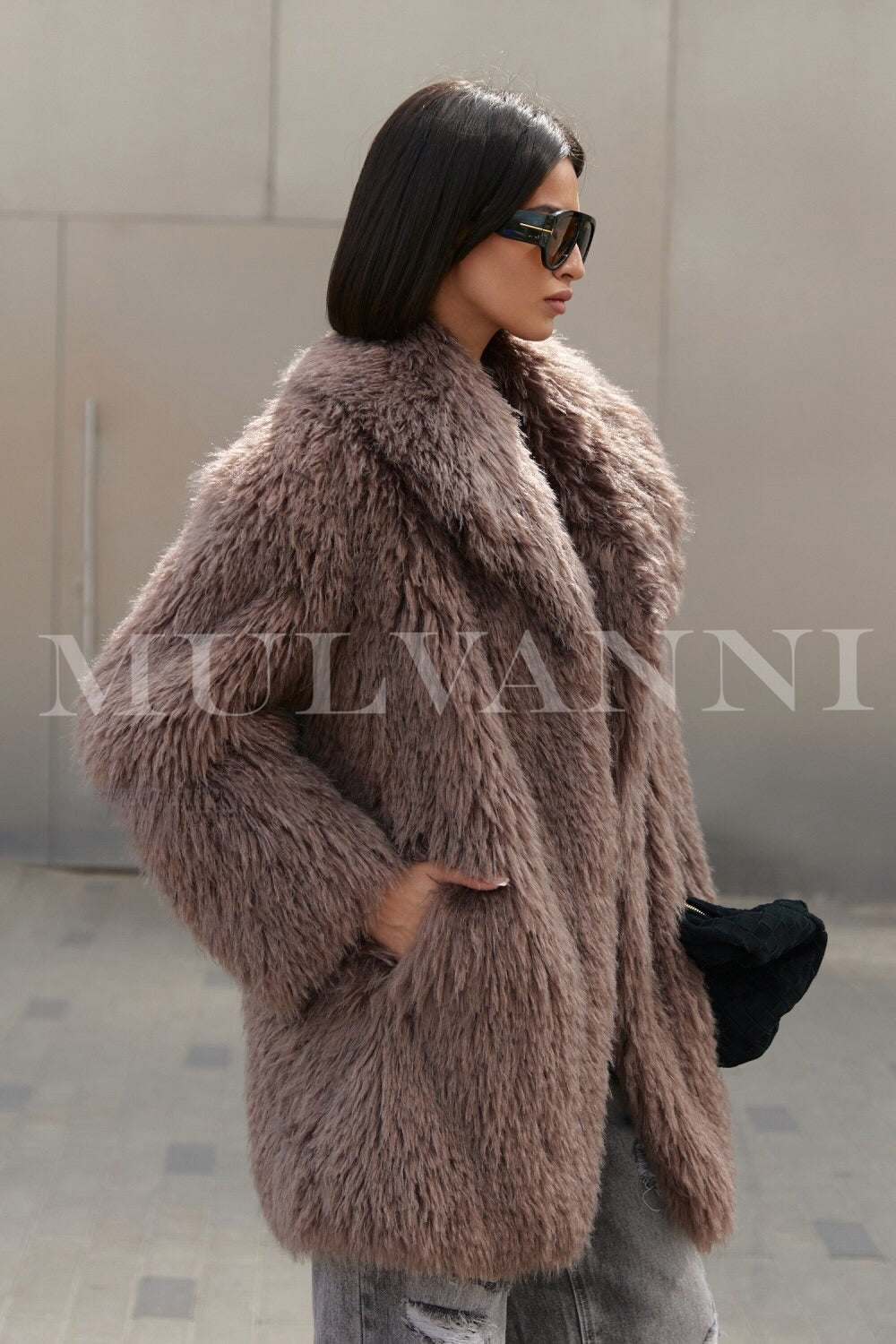 A woman in a brown faux Mongolian coat, showcasing a soft, fluffy texture, complemented by sleek sunglasses. The coat's elegant design adds warmth and style, making it a standout choice for cold weather.