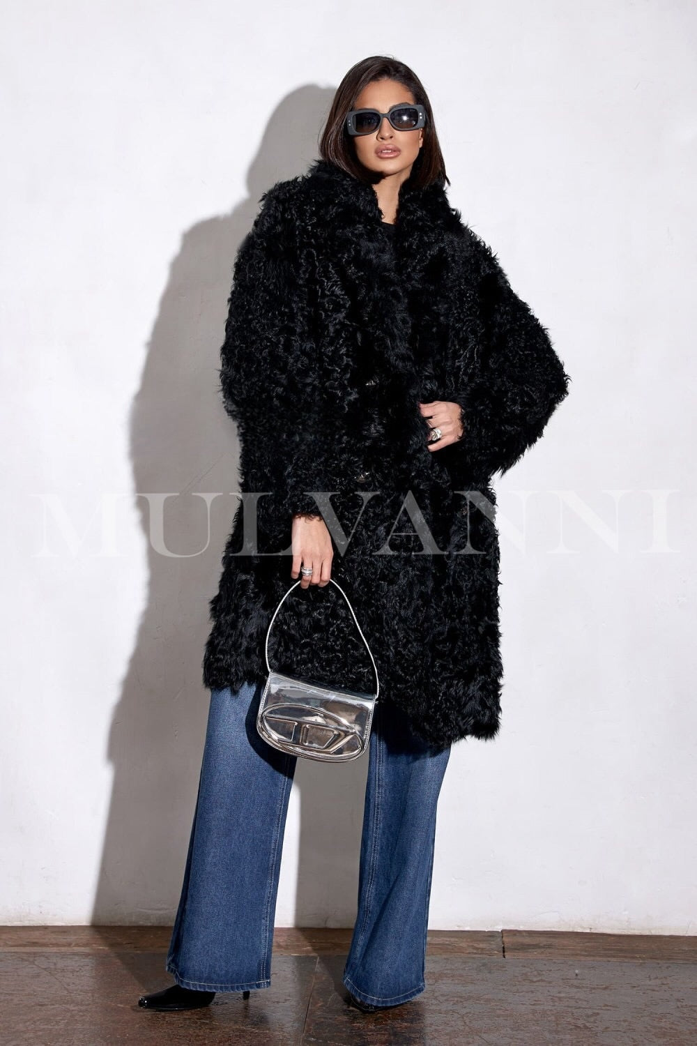 A woman draped in a long black shearling coat, showcasing its rich, fluffy texture and flowing design. The sunglasses add a fashionable flair, creating a chic and polished winter ensemble.