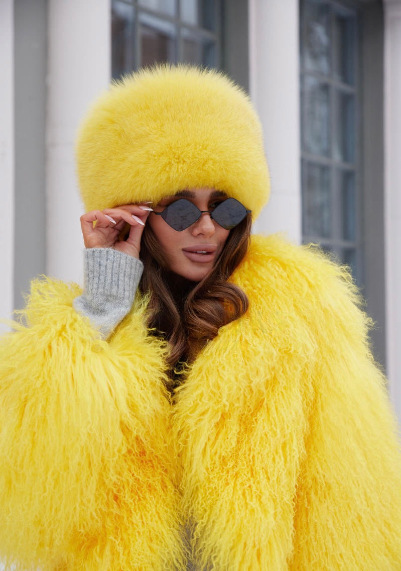 A woman wearing a vibrant yellow fox fur hat paired with a matching Mongolian fur coat. The hat features a plush, textured finish, while the coat showcases a soft, luxurious design, creating a bold and elegant winter look.