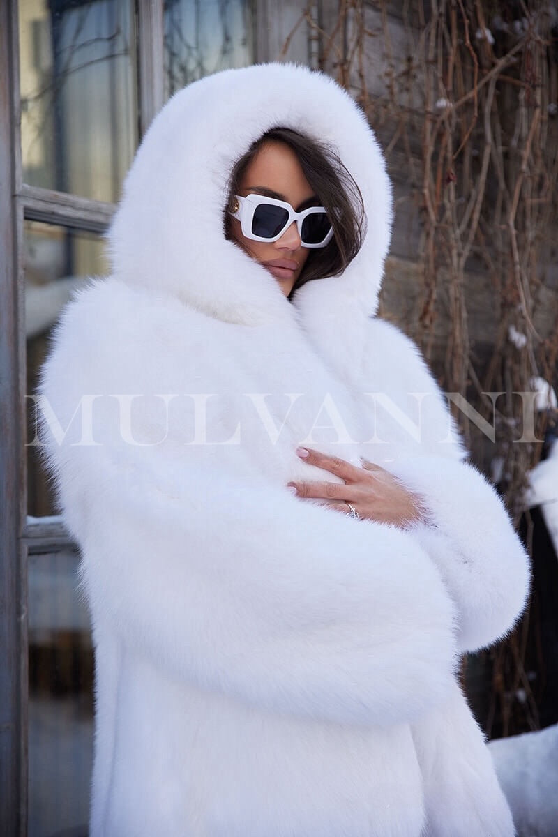 A woman draped in a glamorous white hooded fox fur coat, highlighting its luxurious texture and elegant silhouette. The coat includes a spacious hood and falls gracefully, perfect for a sophisticated winter ensemble.