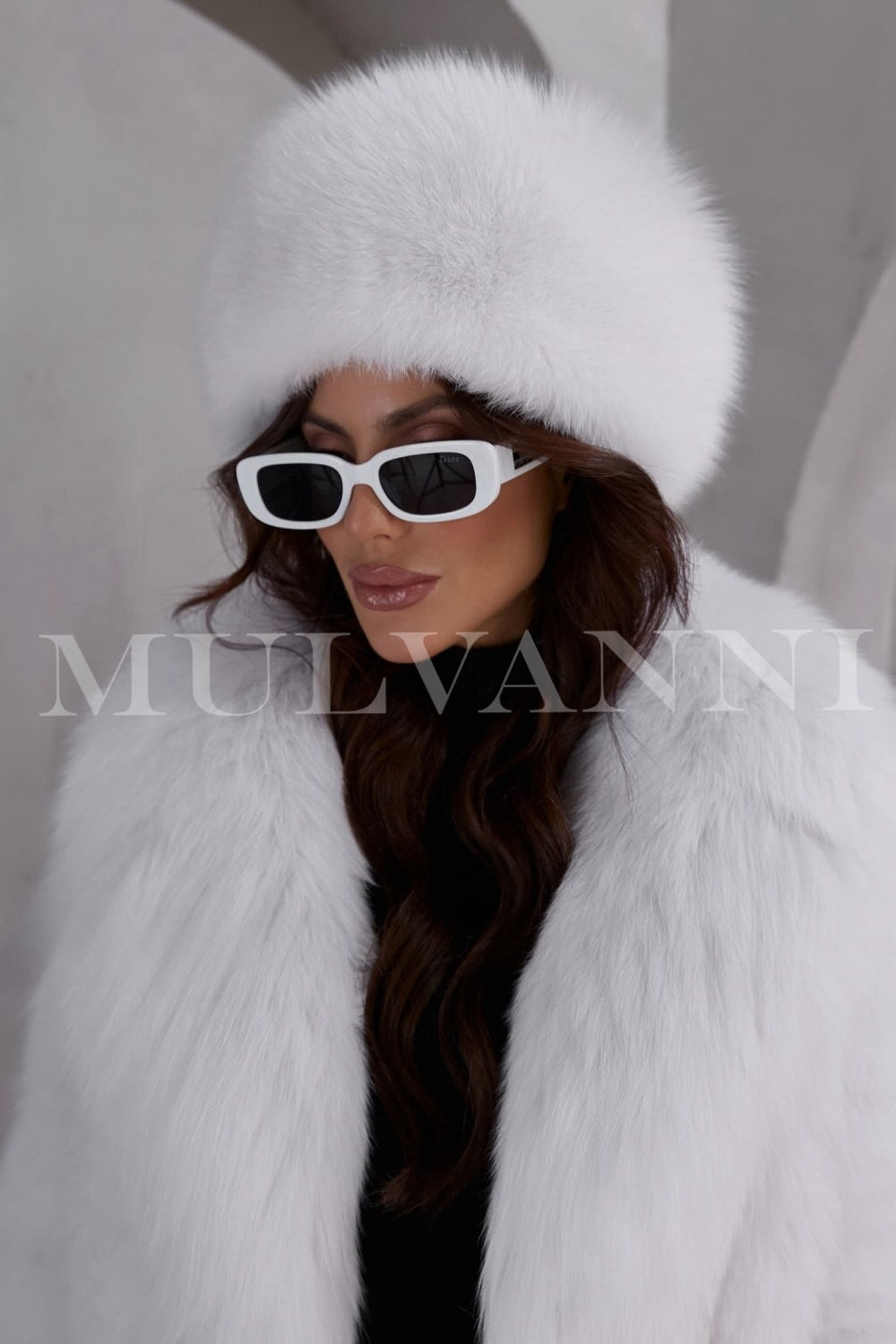 A woman wearing a white fox fur hat, featuring a soft, luxurious texture and a snug fit. The hat has a stylish, rounded design that frames her face, adding an elegant touch to her winter outfit.