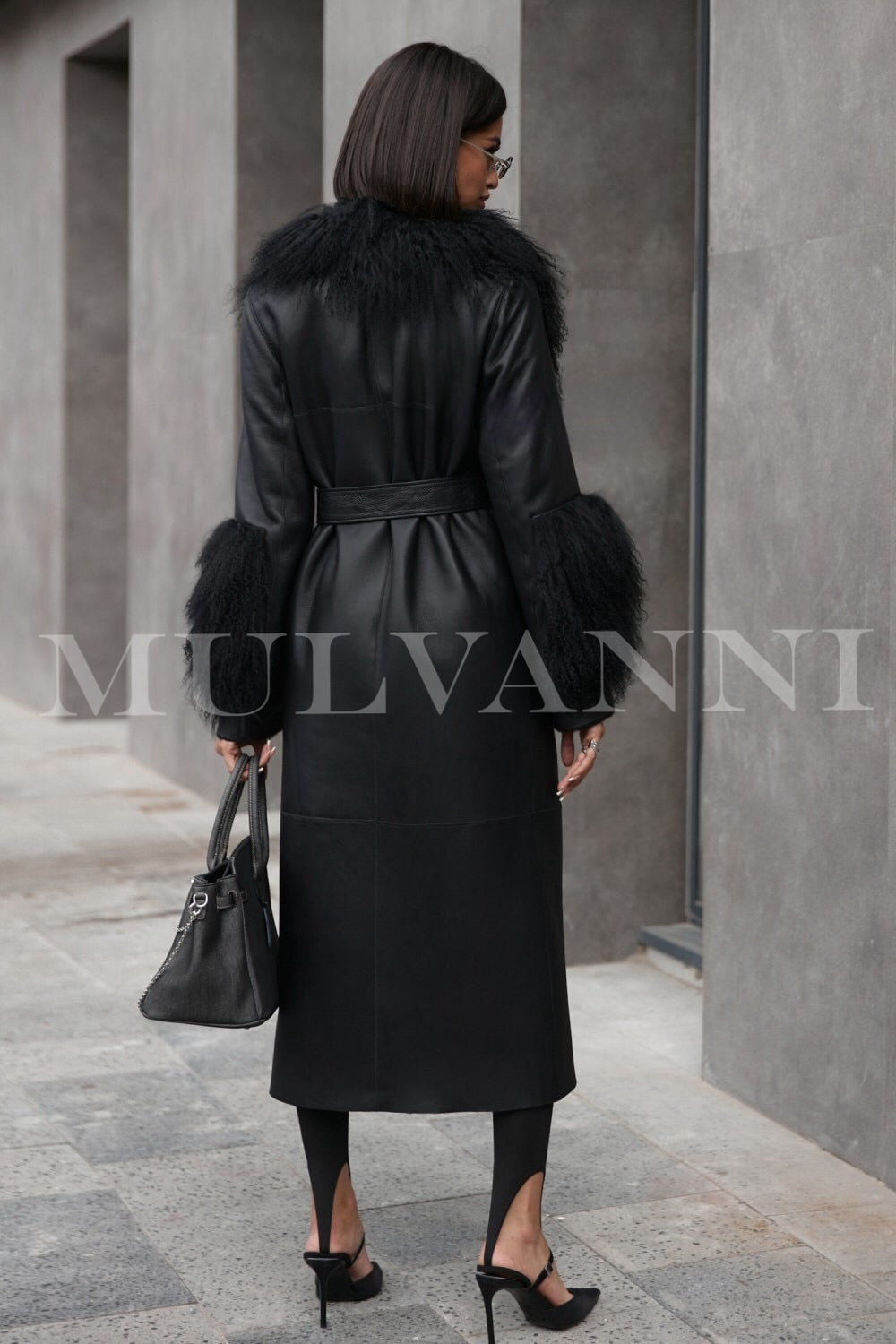 A woman wearing a chic black leather trench coat, enhanced by Mongolian fur detailing on the collar and cuffs. The coat's sleek design and warm materials offer both elegance and comfort for cold seasons.