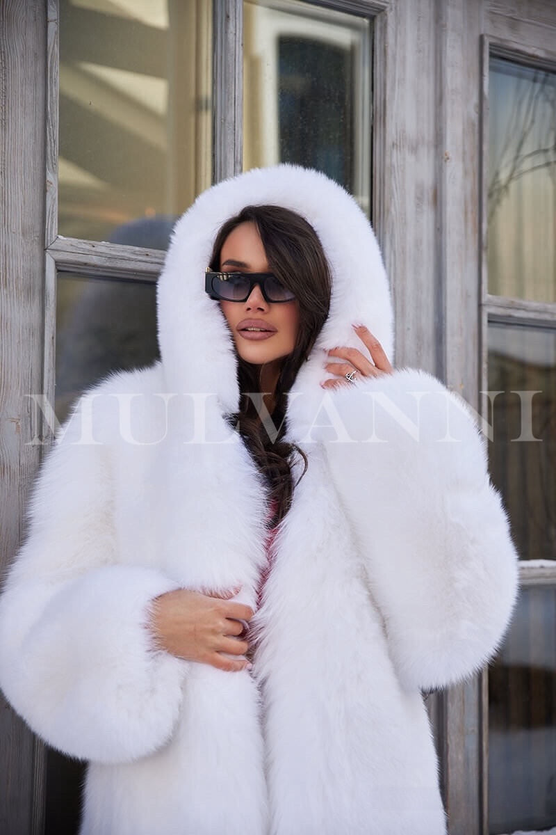 A woman in an elegant white hooded fox fur coat, featuring a soft, plush exterior and a cozy hood. The coat has a flattering design, making it a perfect statement piece for winter fashion.