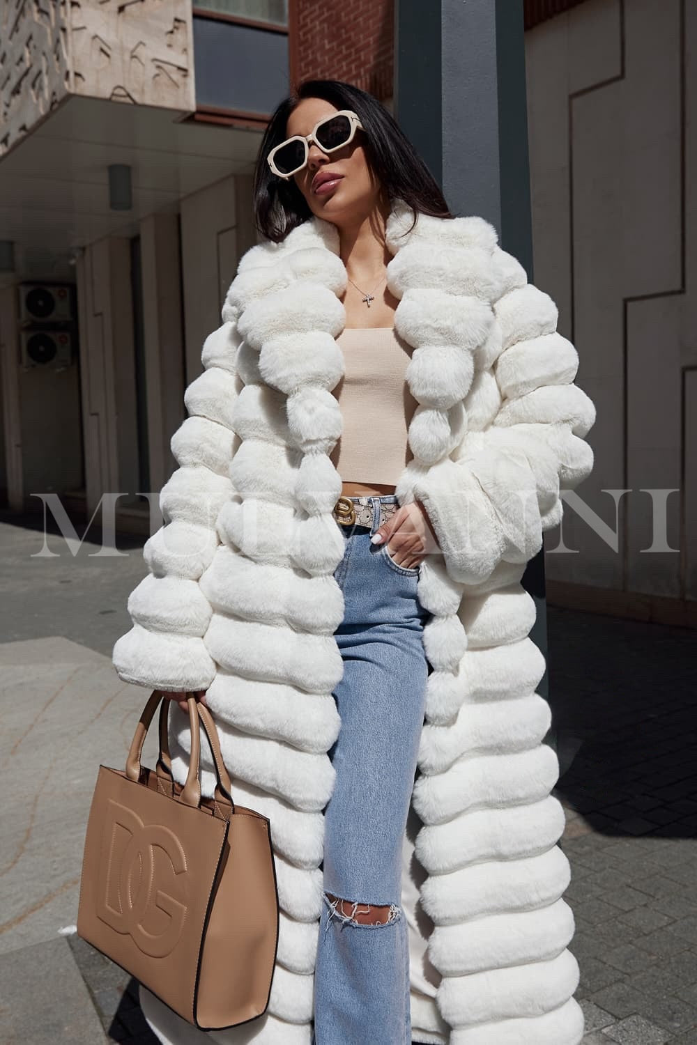 A woman wearing a long white faux fur coat, featuring a soft, high-quality texture and a matching belt that cinches the waist for a flattering fit. The coat exudes elegance, combining luxurious style with a sophisticated silhouette.