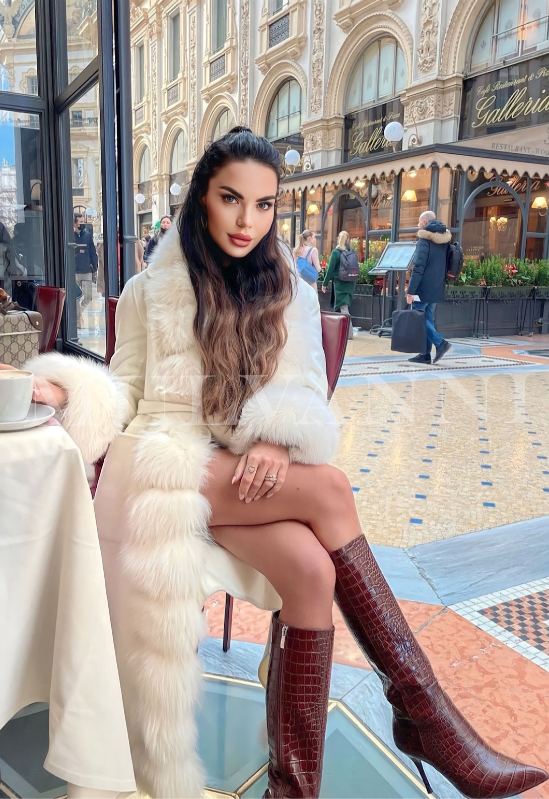 Elegant woman wearing ALIZE white cashmere coat with fox fur trim, sitting outdoors at a chic cafe in a stylish city setting.