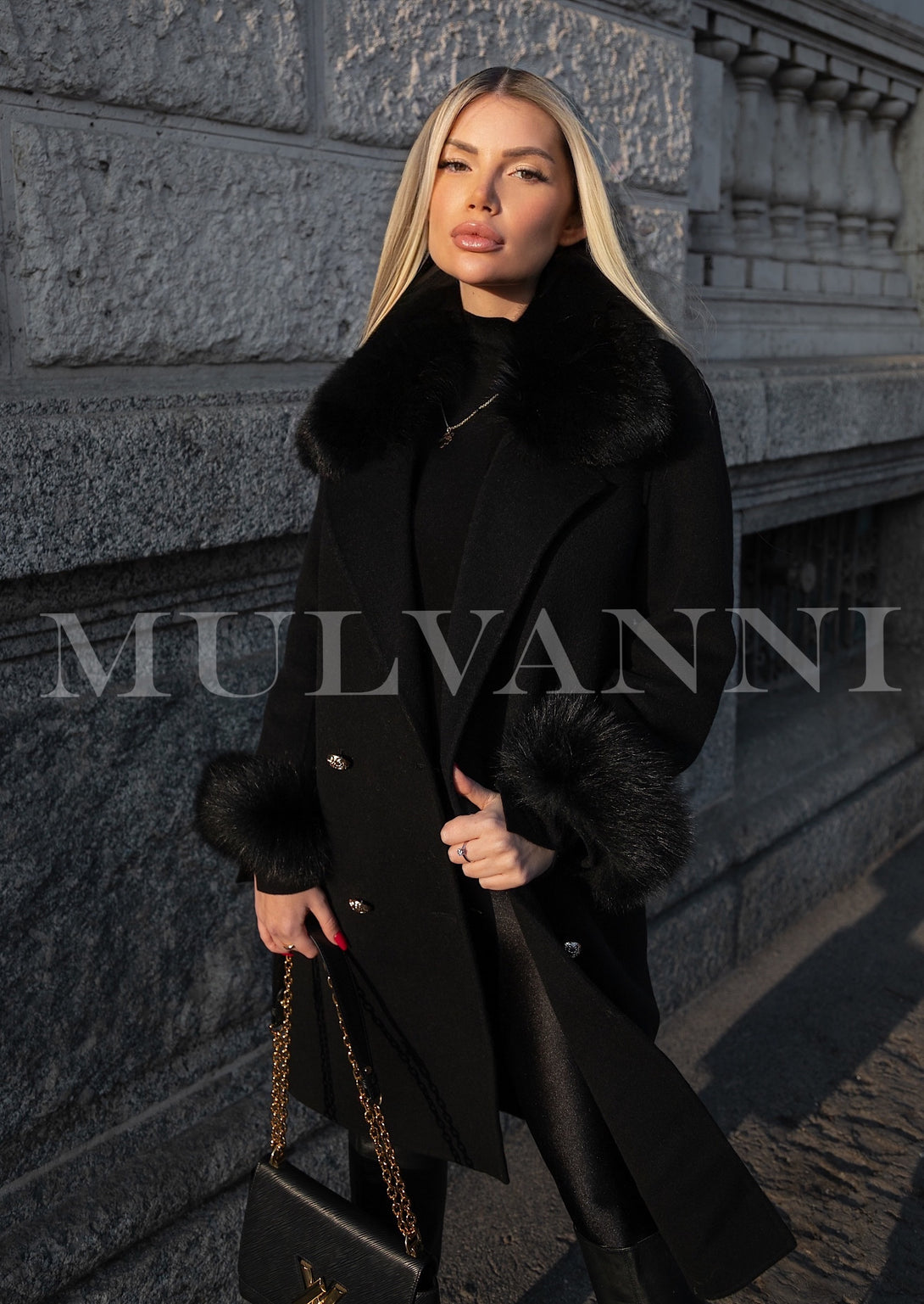 A woman dressed in a chic black cashmere coat with soft fox fur trim, exuding sophistication and warmth. The coat's refined design makes it a standout piece for cold-weather occasions.